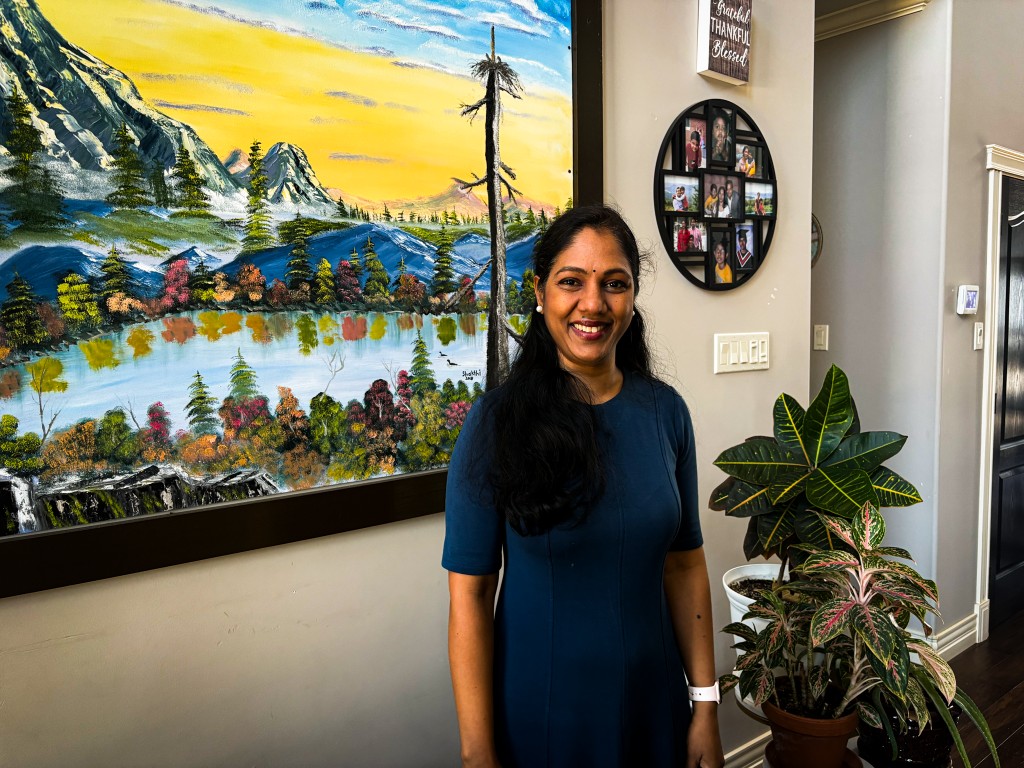 A woman wearing blue dress smiles and stands infront of a painting on a wall.